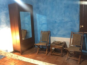 Een kamer met een blauwe wand, twee rieten stoelen, een klein tafeltje met een schaal en een kaars, en een hoge kast.