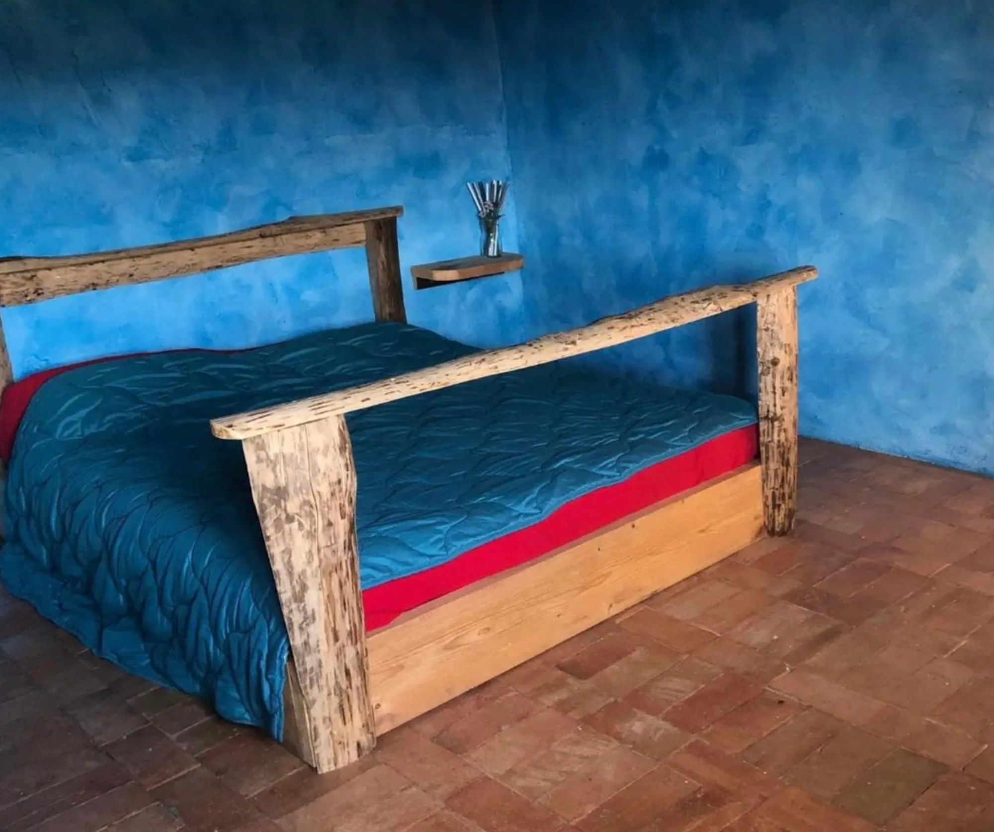 Een houten bed met een matras in rood en blauw in een kamer met blauwe wanden en een vloer van baksteen.