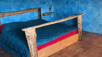 Een houten bed met een matras in rood en blauw in een kamer met blauwe wanden en een vloer van baksteen.