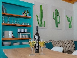 Ein Essbereich mit einem Tisch für zwei Personen, einer Flasche El Greco Wein und zwei Weingläsern. Regale mit Gläsern und Geschirr, sowie eine Wand mit Kakteendekorationen.