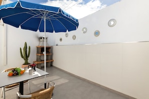 Um pátio com mesa, cadeiras, um guarda-sol azul, um cacto, churrasqueira e arte decorativa na parede.