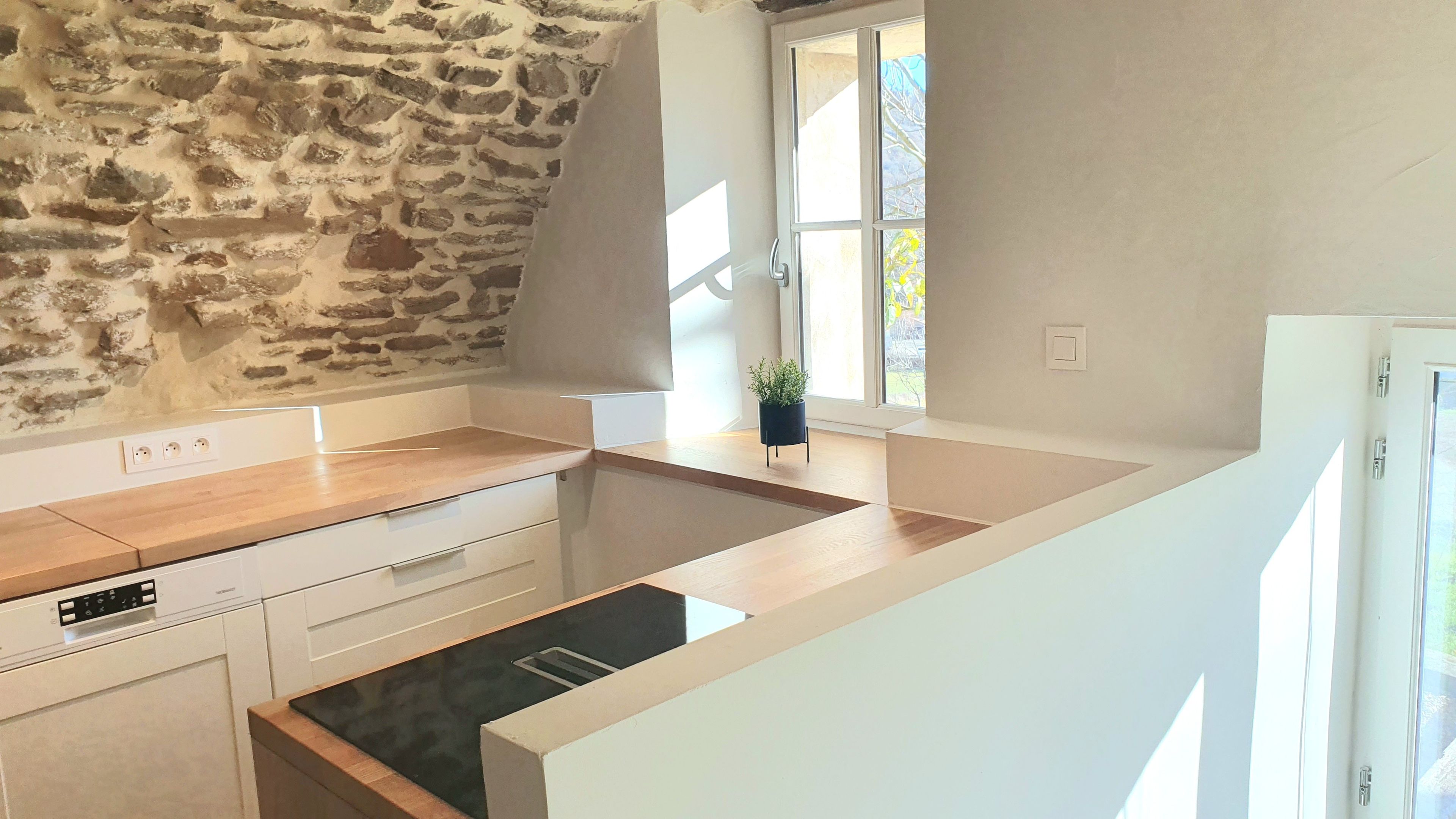 Cozinha com parede de pedra, bancada de madeira, armários brancos e janela com vista.