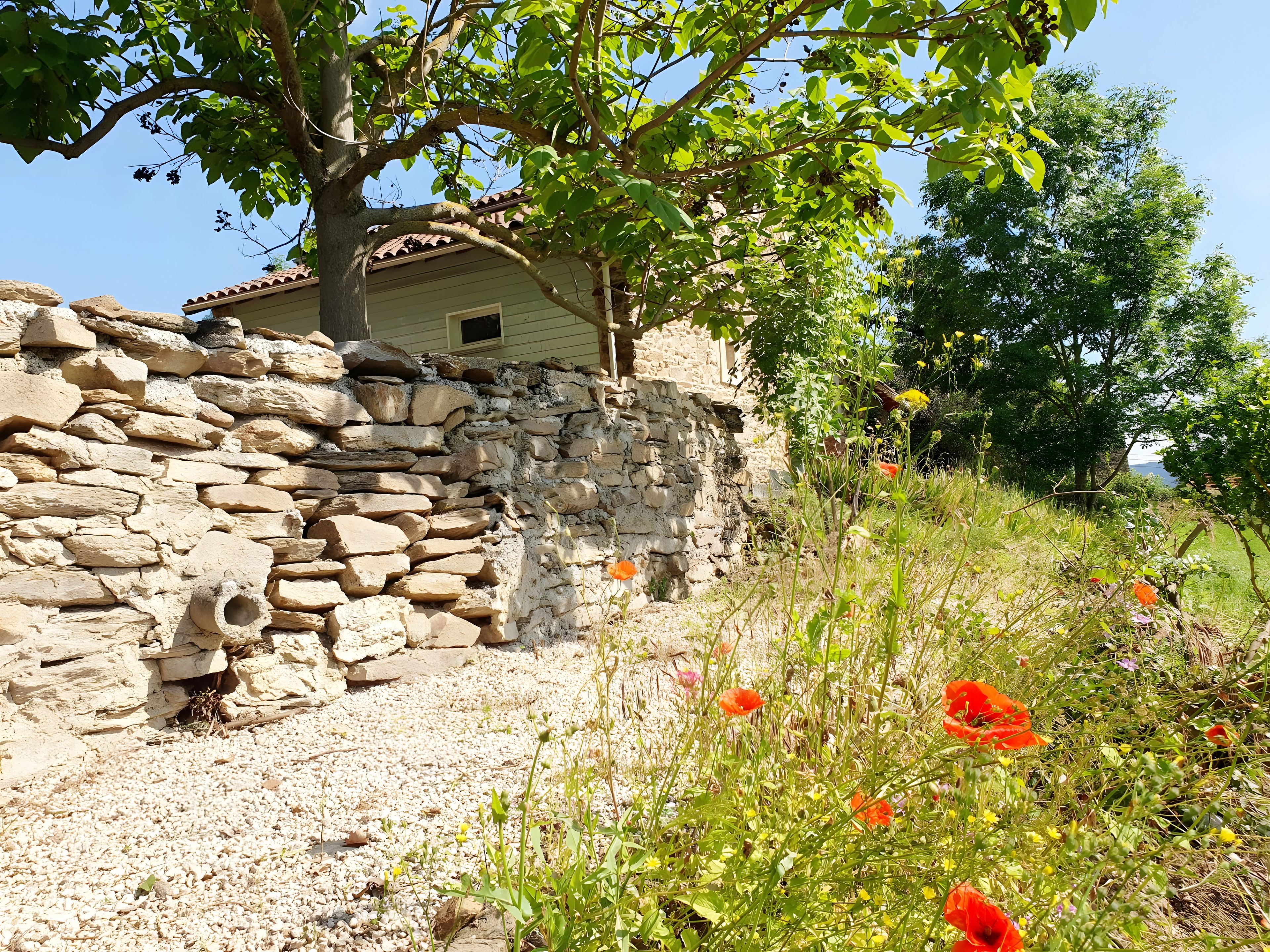 Un mur de pierre, avec une maison de style rustique qui se devine en partie derrière, le tout entouré de verdure et de fleurs sauvages.