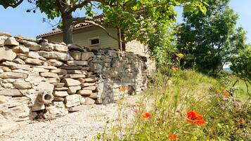 Un mur de pierre, avec une maison de style rustique qui se devine en partie derrière, le tout entouré de verdure et de fleurs sauvages.