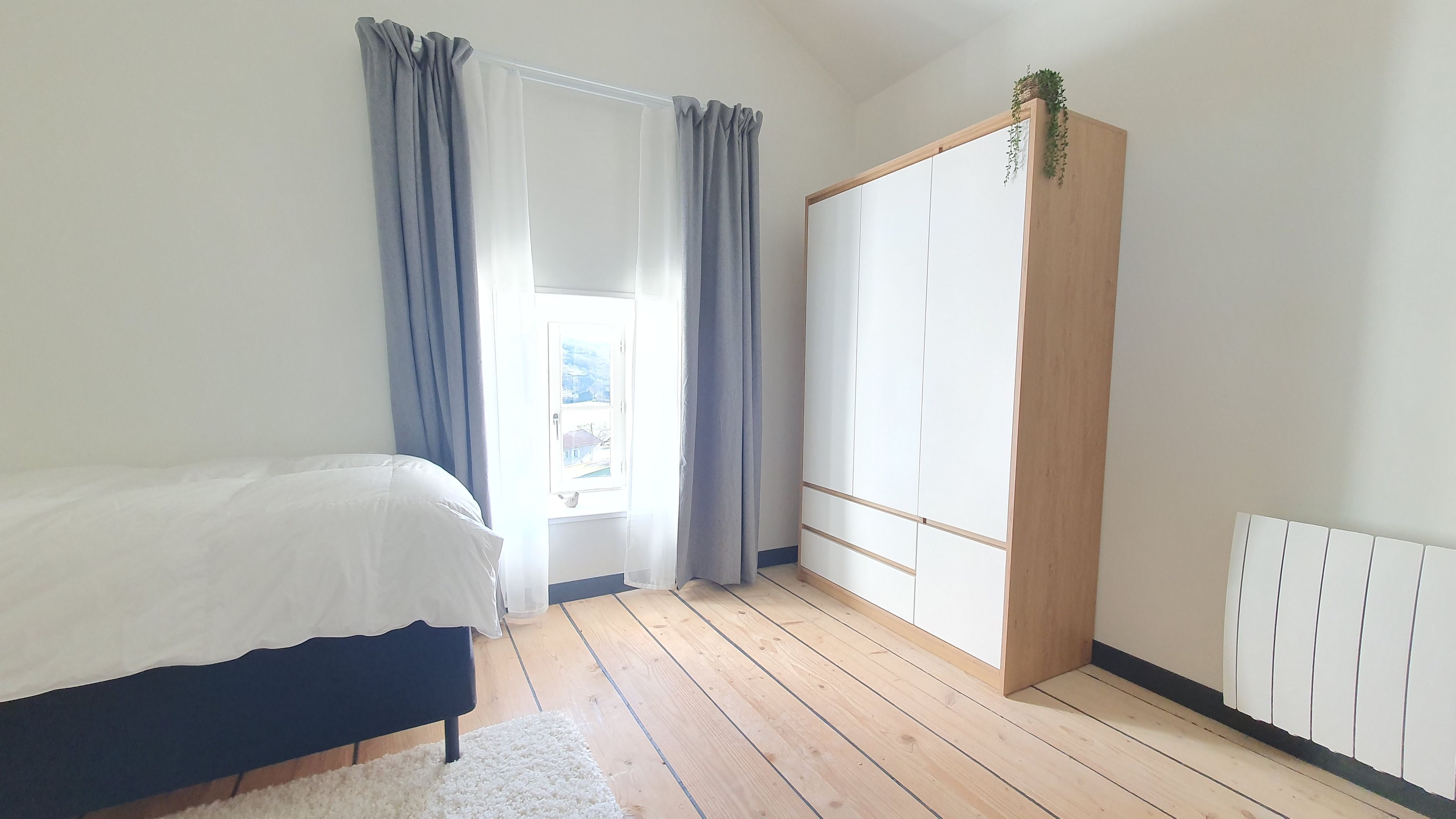 A bedroom with a bed, a wardrobe, a window with curtains, and a radiator.