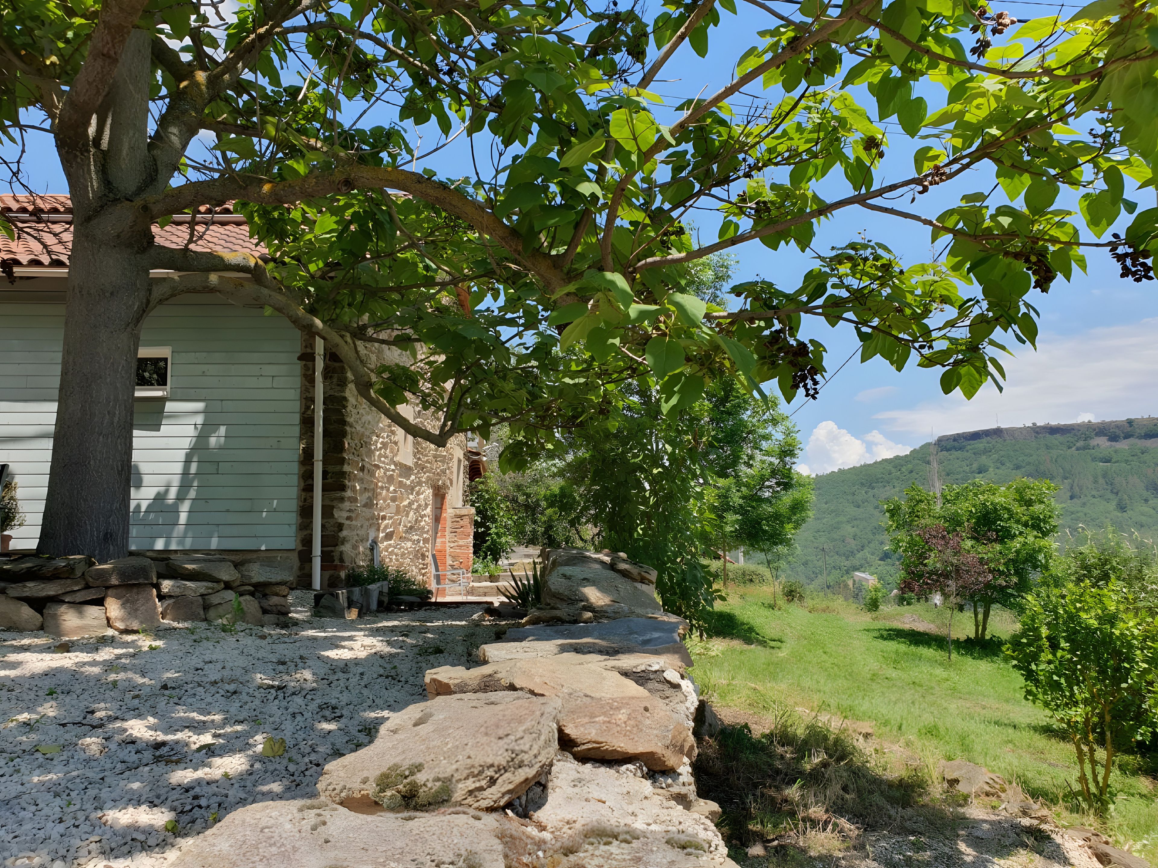 A rustic stone building with a sloped roof, surrounded by a gravel path and greenery.