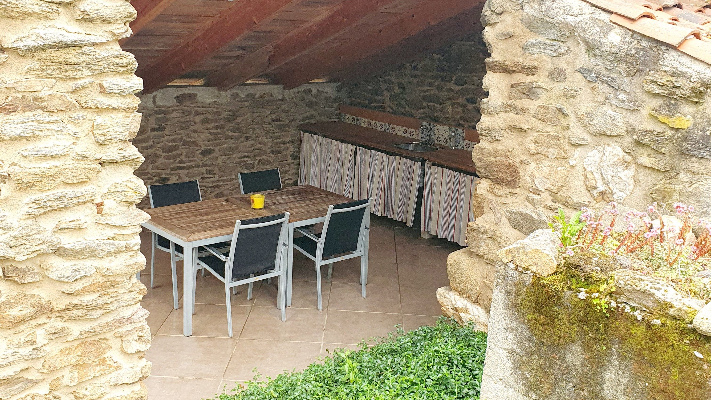 A stone-walled outdoor dining area with a wooden table and chairs, a tiled floor, and a stone wall with vegetation.