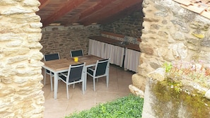 A stone-walled outdoor dining area with a wooden table and chairs, a tiled floor, and a stone wall with vegetation. - Holiday Home 'Le Poulailler' with Private Garden, Summer Kitchen and Stunning Views (Blesle)