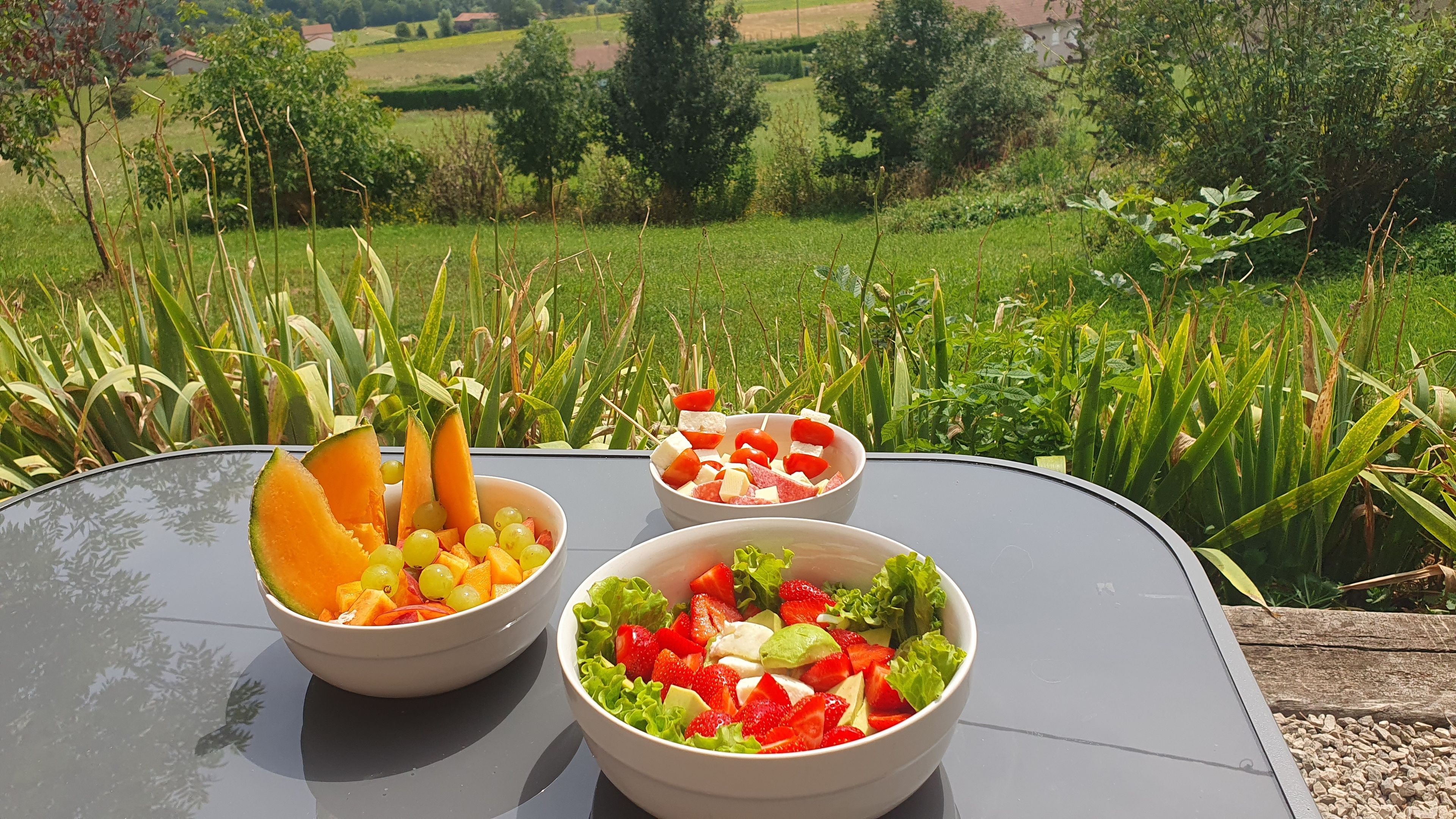 A table with three bowls of fruit salad, set outdoors with a view of a green landscape.