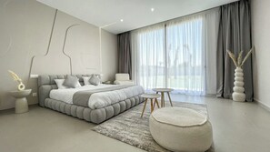 Un dormitorio moderno con una cama grande, mesitas de noche, un otoman redondo y vistas al exterior a través de cortinas translúcidas.