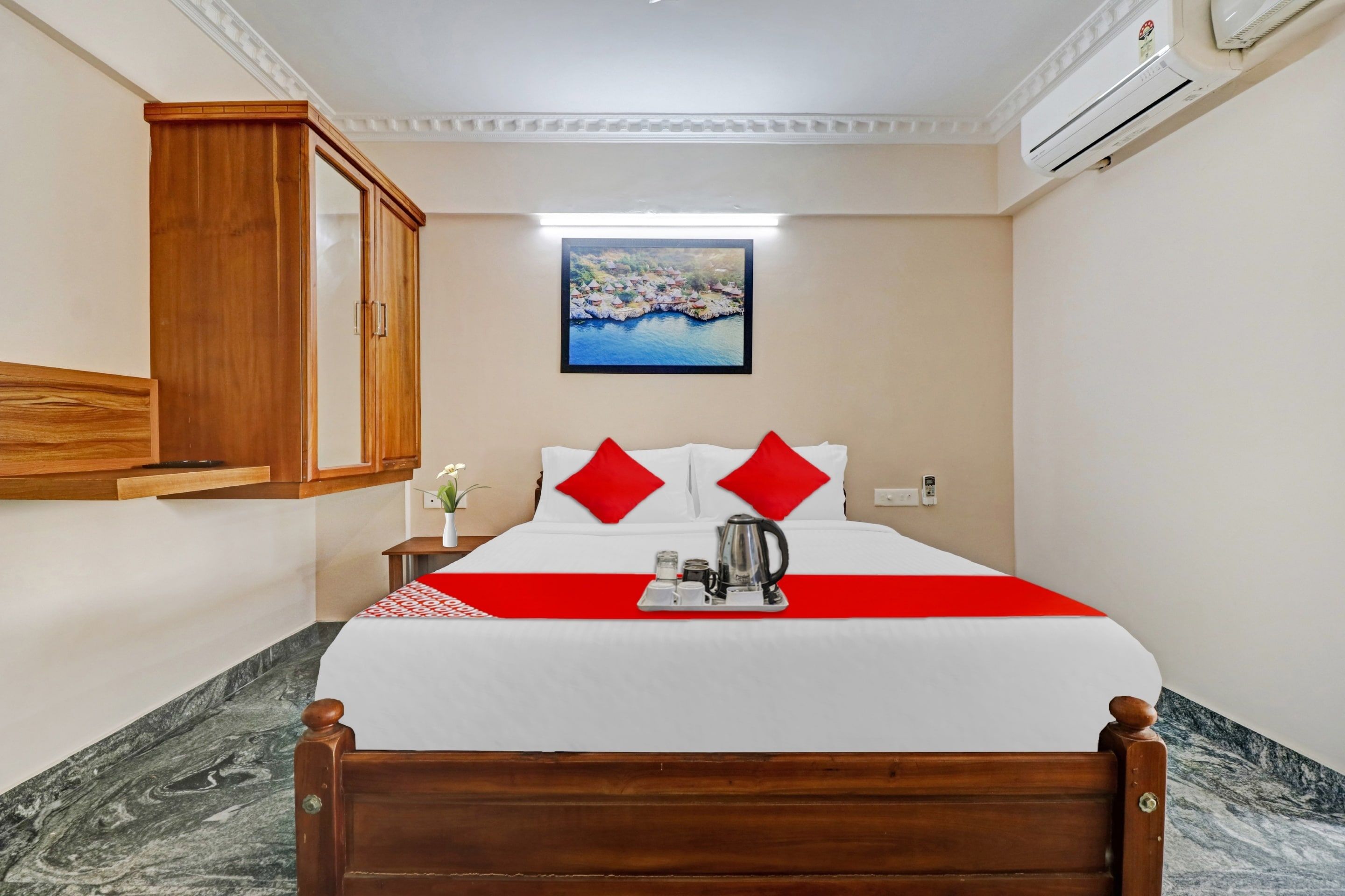 Een hotelkamer met een bed, nachtkastje, een rood-wit sprei, een schilderij aan de muur en een airconditioning.