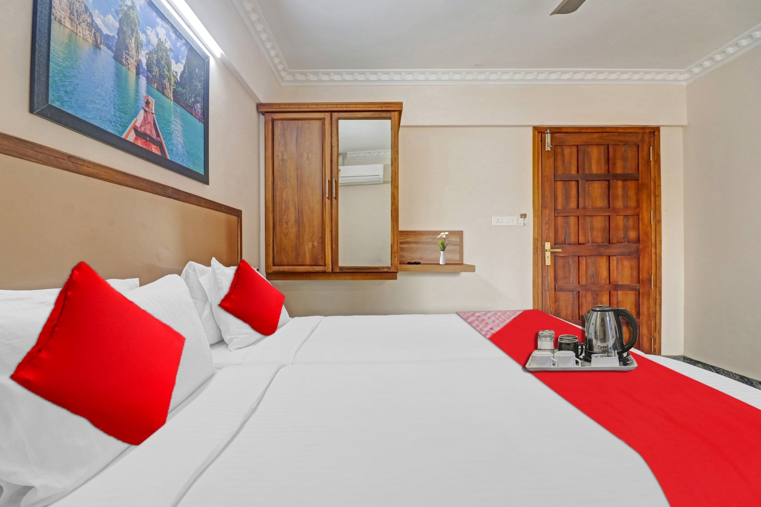 Habitació d'hotel amb un llit, un llençol escacat vermell i blanc, un armari de fusta, un mirall, un quadre a la paret i una safata amb una paperera i gots.