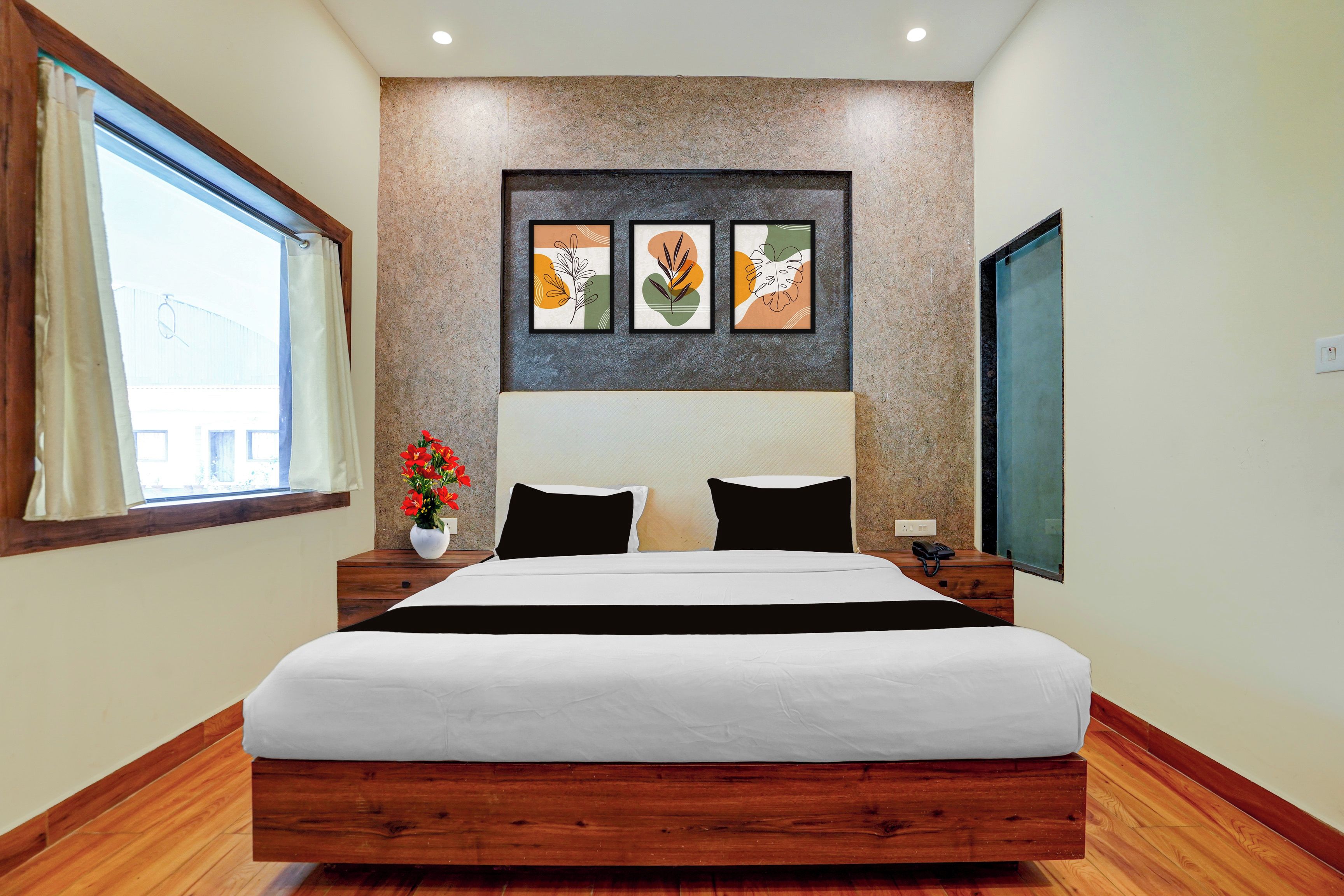 A hotel room with a large bed, a wooden headboard, a window with curtains, a wall-mounted artwork, and a bedside table with a vase of flowers.