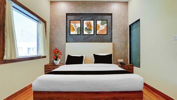 A hotel room with a large bed, a wooden headboard, a window with curtains, a wall-mounted artwork, and a bedside table with a vase of flowers.