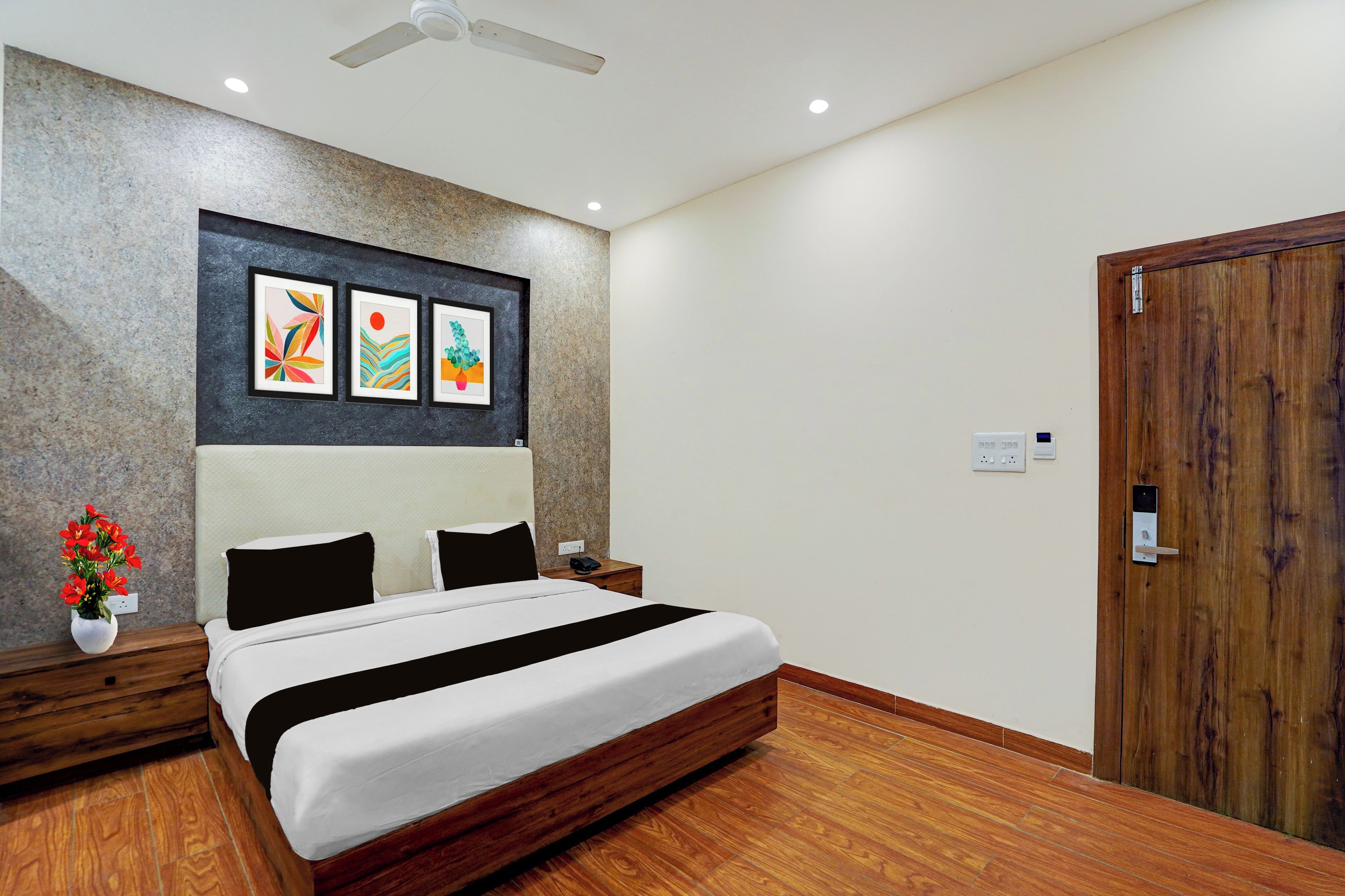 A hotel room with a bed, a nightstand, a wall-mounted artwork, a wooden door, and a ceiling fan.