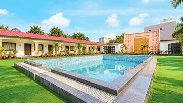A swimming pool with a tiled deck, surrounded by a well-maintained lawn and a row of buildings with red roofs.