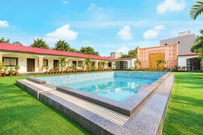 A swimming pool with a tiled deck, surrounded by a well-maintained lawn and a row of buildings with red roofs. - Townhouse Oak Rapti Riverview Formerly (Gorakhpur)