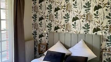 Ein Schlafzimmer mit einem Bett, einem Kopfteil, einem Fenster mit Vorhängen und einer Wand mit einem Blattmuster.