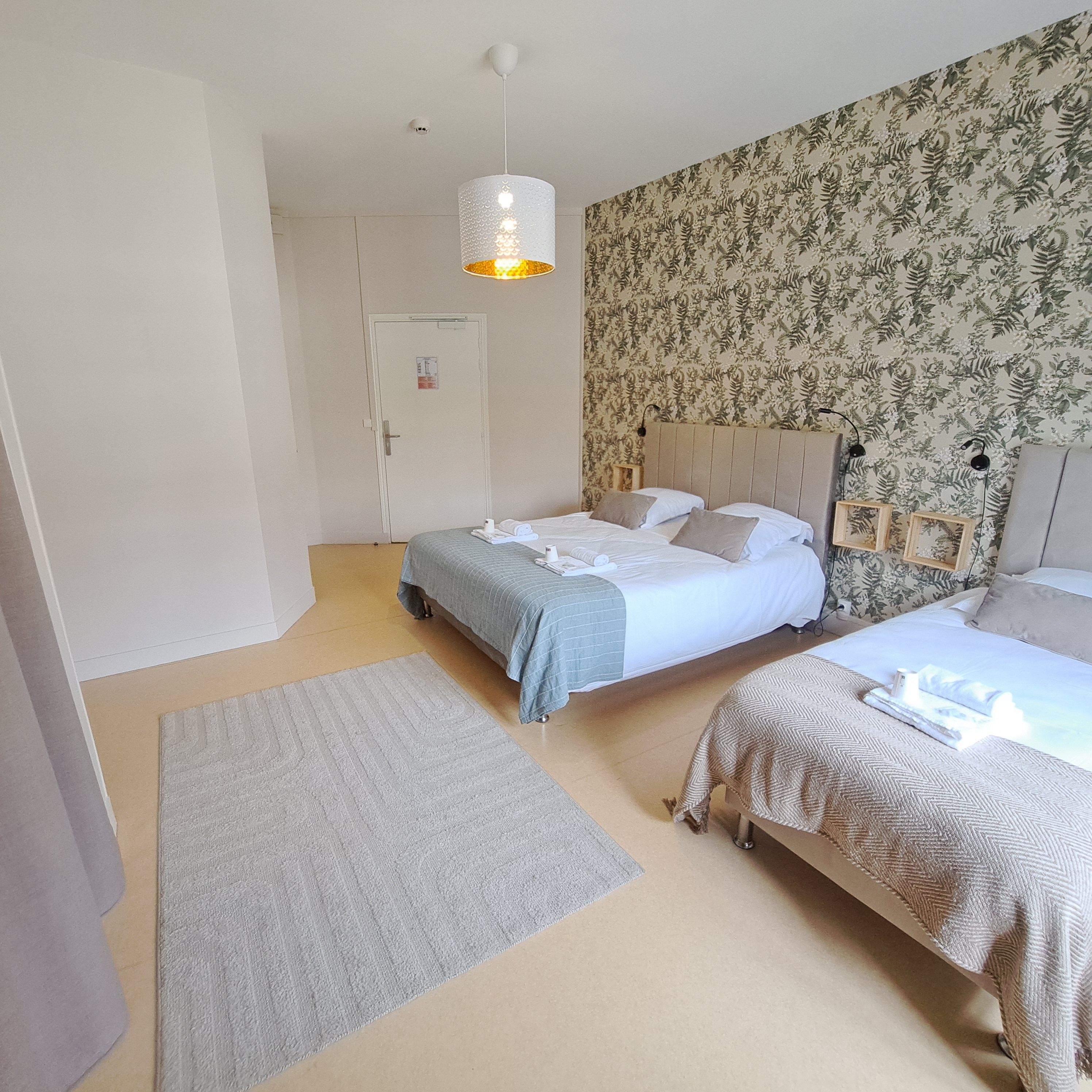 Ein Hotelzimmer mit zwei Betten, einem grauen Teppich, blumigem Wandpapier und einer Pendelleuchte.
