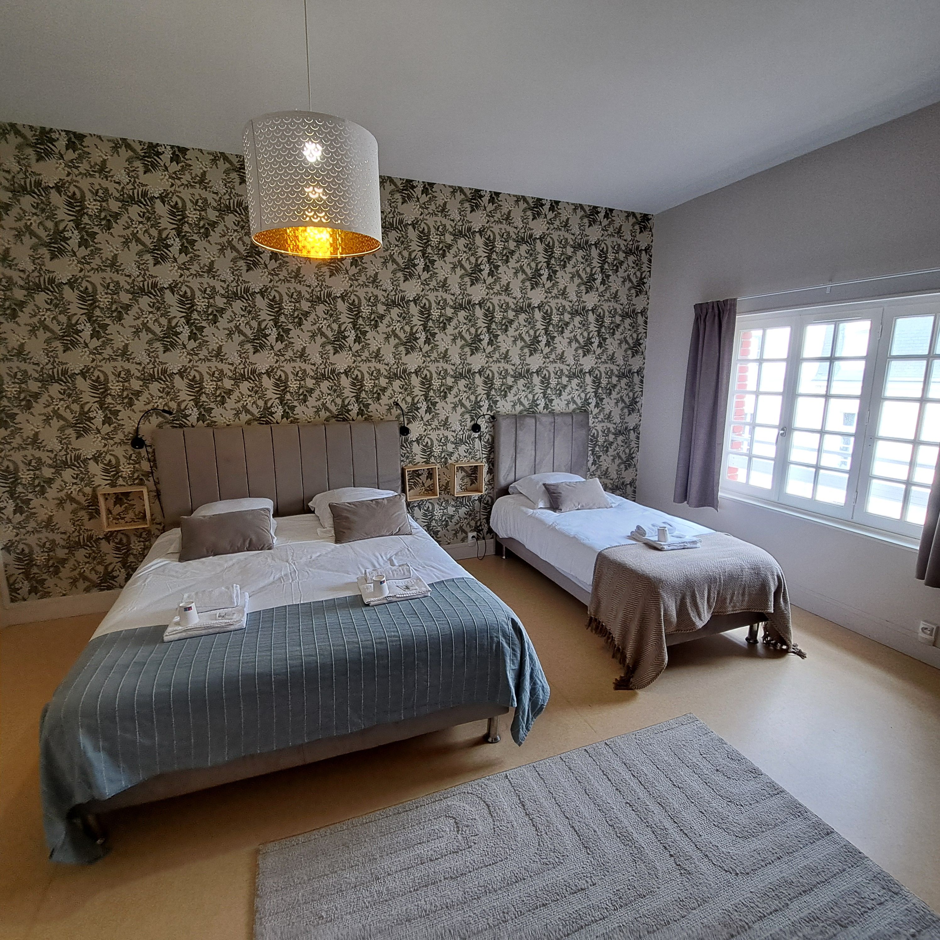 A hotel room with two beds, a patterned wallpaper, a hanging light fixture, and a window with curtains.