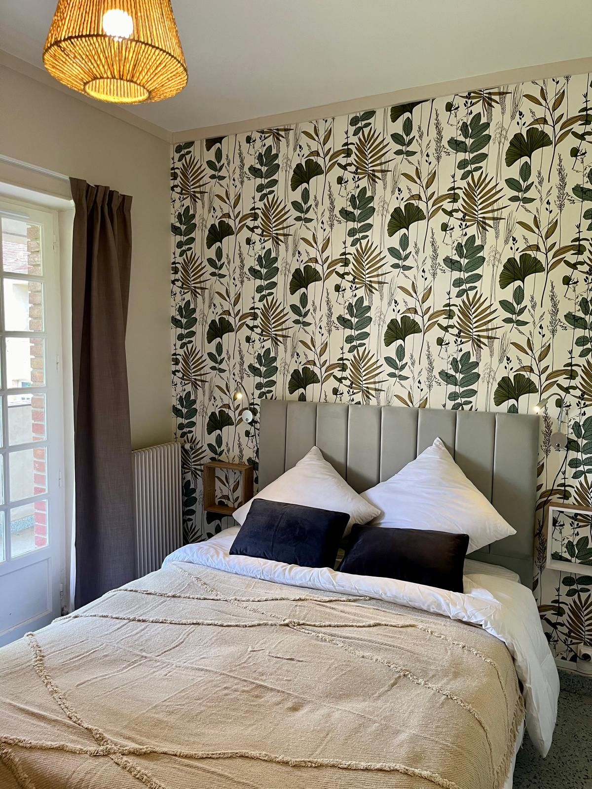 Een slaapkamer met een bed, hoofdbord, raam met gordijnen en een muur met bladmotief.