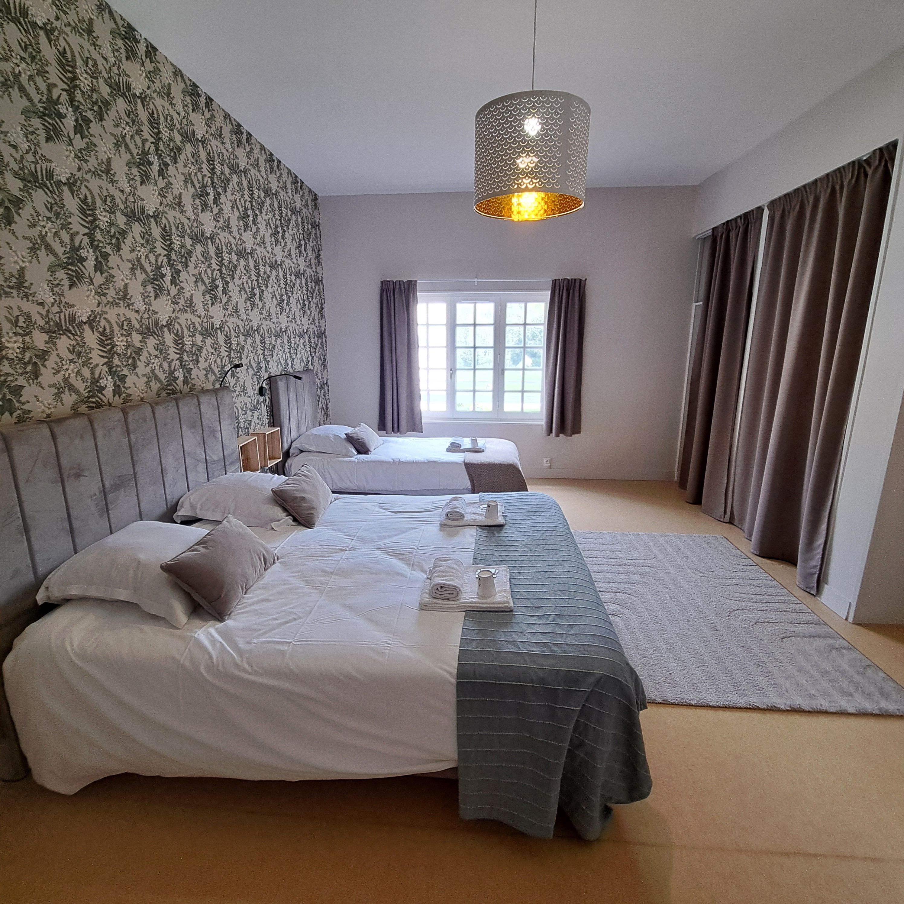 A hotel room with two beds, a patterned wallpaper, a hanging light fixture, a window with curtains, and a carpeted floor.