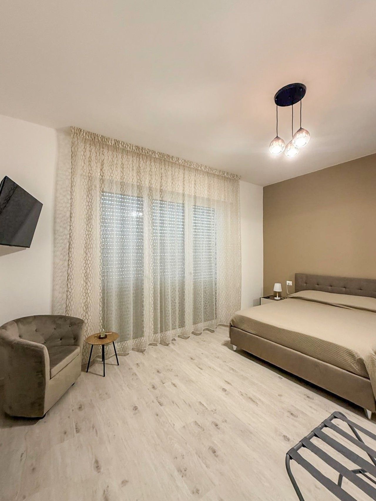 Een slaapkamer met een bed, een stoel, een klein tafeltje, een aan de muur bevestigde televisie en een plafondlamp.
