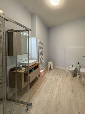 Un baño moderno con cabina de ducha de vidrio, tocador de madera, lavamanos blanco, un bidé y un inodoro.
