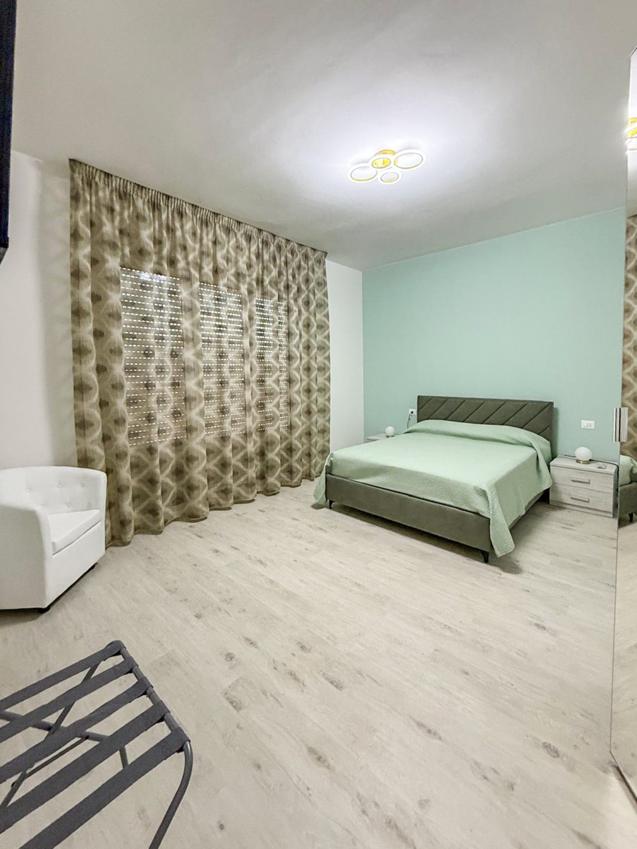 Ein Schlafzimmer mit einem Bett, einem Nachttisch, einem Sessel und einem gemusterten Vorhang.