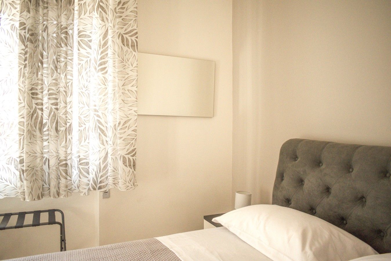 Ein Hotelzimmer mit einem Bett, einem Nachttisch, einer Lampe und einem Fenster mit einem gardiniertem Vorhang mit Blattmuster.