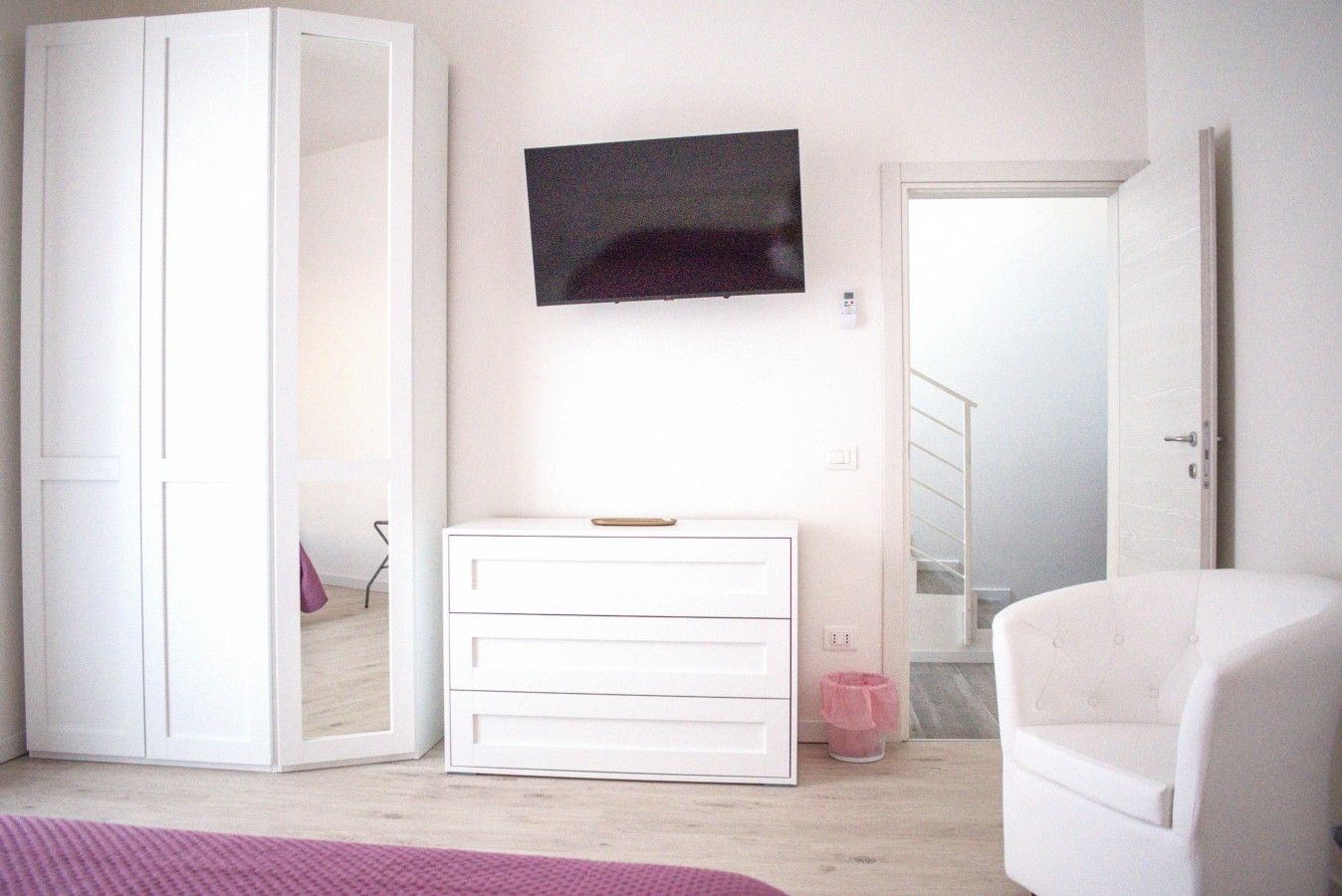 Une chambre avec une armoire blanche, une table de chevet, un téléviseur fixé au mur et une couvre-lit rose.