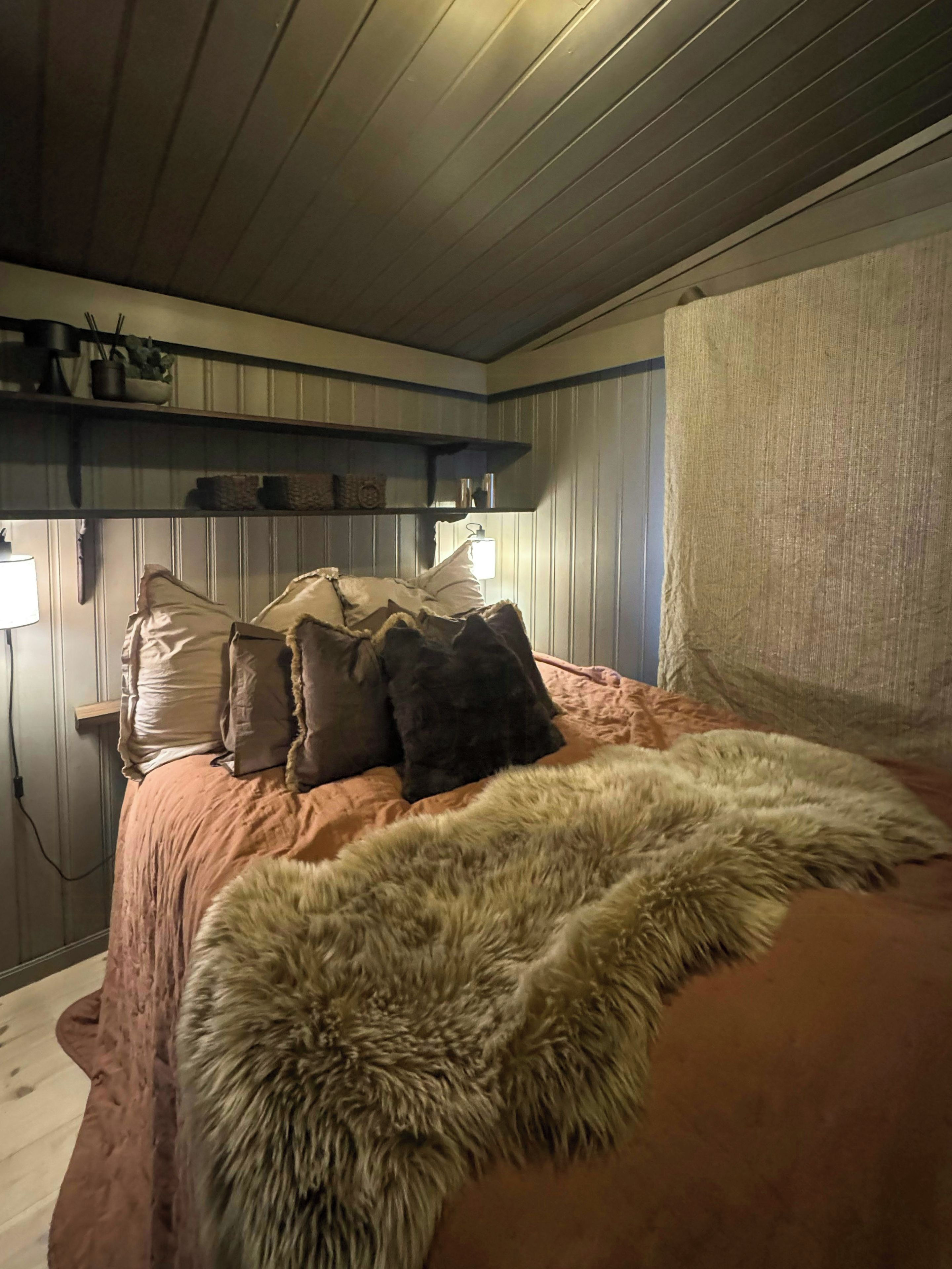 Ein Schlafzimmer mit einem Bett, Kissen, einem Fellüberwurf, einem Regal mit Gegenständen und einer Wandleuchte.