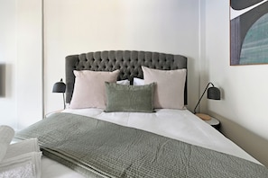 Un lit bien fait, avec une couverture grise, une tête de lit capitonnée et deux lampes de chevet.