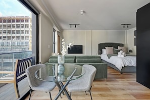 Een moderne slaapkamer met een bed, een bank, een glazen tafel met stoelen en uitzicht op een bouwterrein door een groot raam.