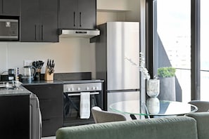 Une cuisine moderne avec des meubles de couleur sombre, une cuisinière en acier inoxydable et un réfrigérateur. On y trouve un coin repas avec une table en verre et une chaise, ainsi qu'un vase rempli de fleurs blanches posé sur le rebord de la fenêtre.