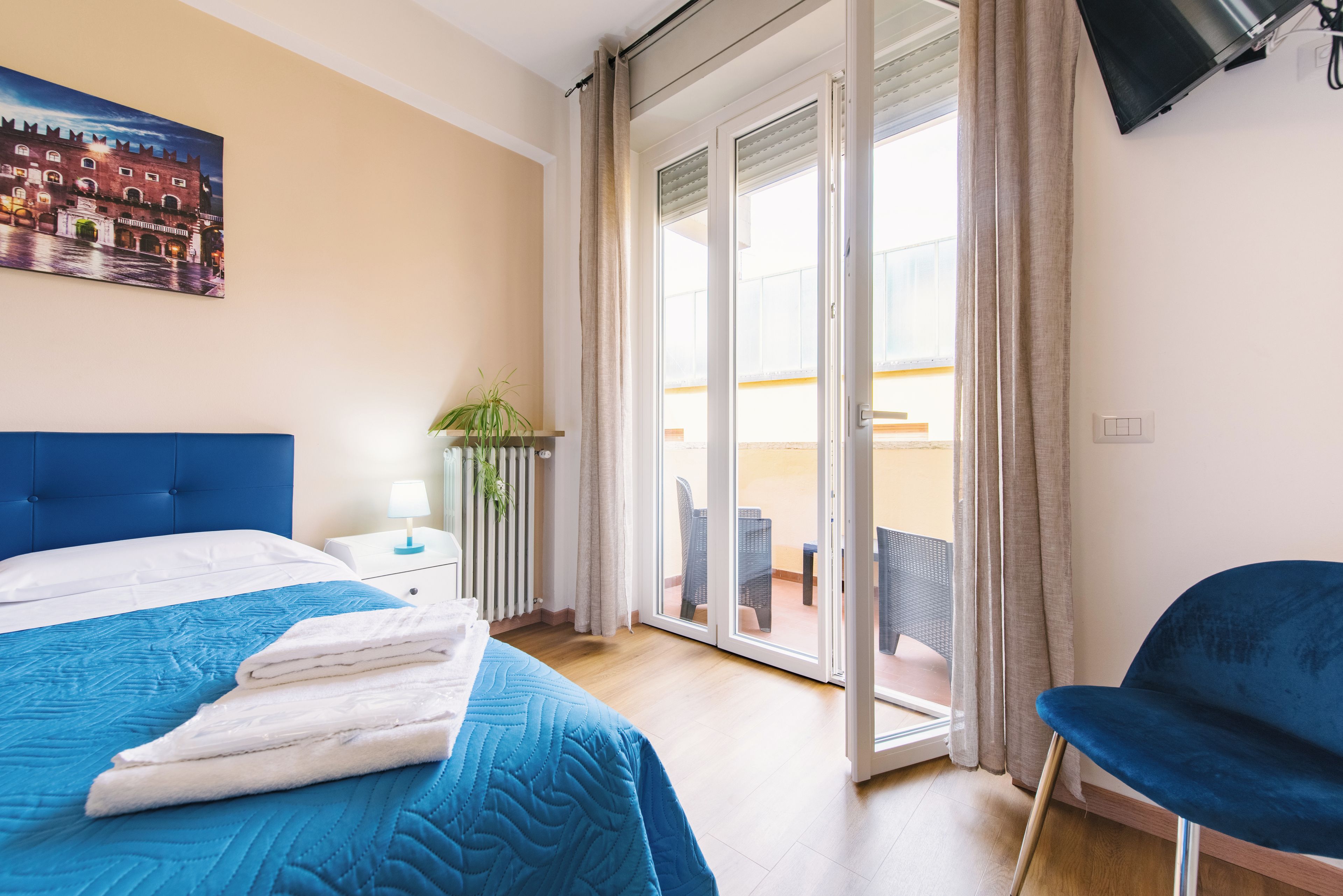 Hotellihuone, jossa on sänky, tuoli, lamppuvarustettu pöytä, kasvi ja parveke, jossa on tuoleja.
