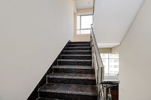 Una escalera con peldaños de mármol oscuro y barandilla de metal, que conduce a un rellano con una ventana.