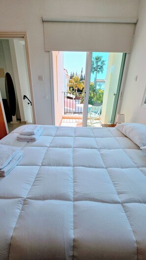 Une chambre d’hôtel avec un grand lit, un balcon donnant sur des palmiers et des immeubles, et une porte qui mène à un couloir.