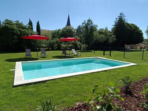 Una piscina rectangular con borde de azulejos, rodeada por un césped. Hay cuatro sillas de descanso y dos sombrillas rojas.