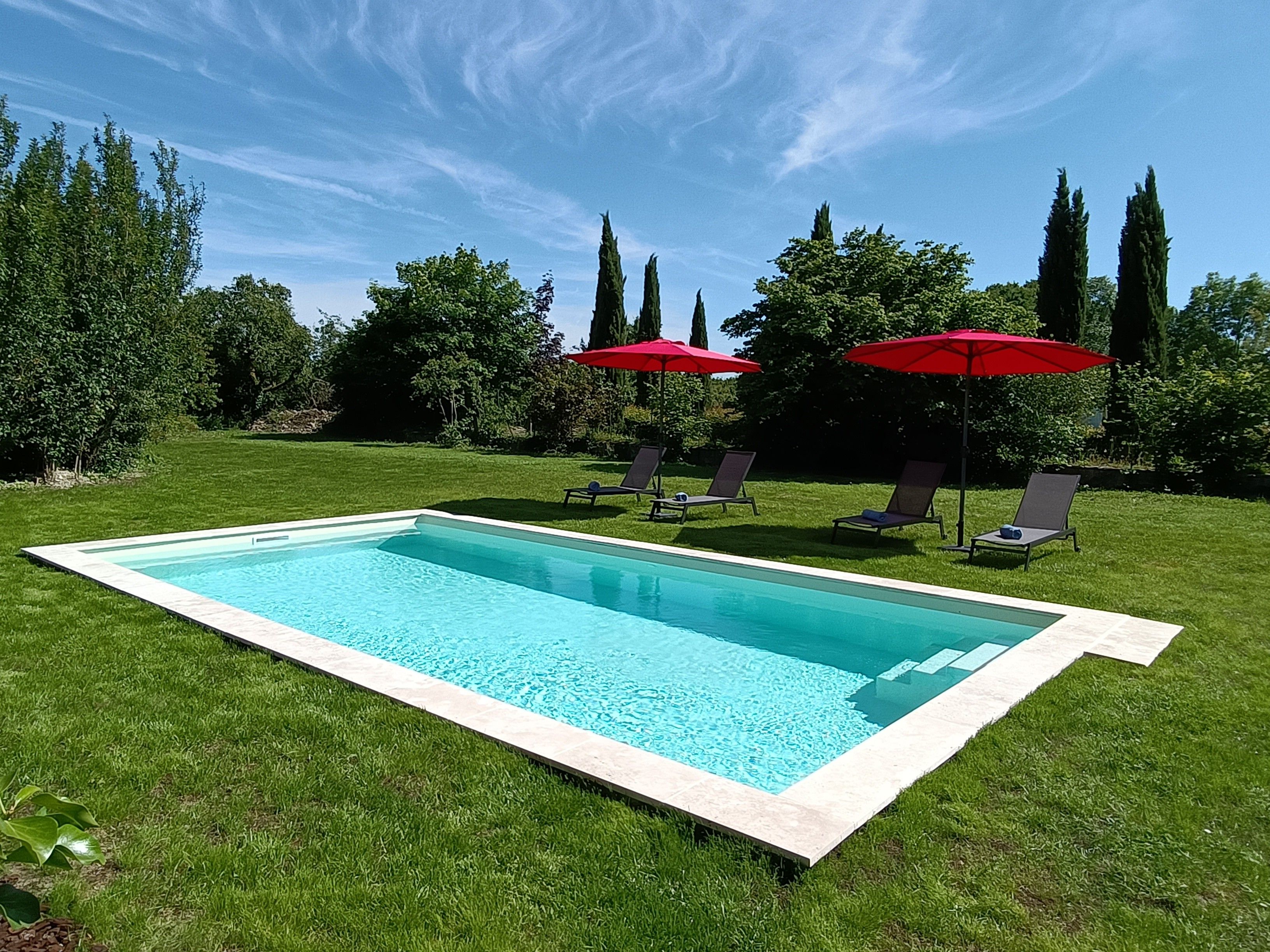 Una piscina rectangular con borde enlosetado, rodeada por un césped bien cuidado y sombrillas rojas.