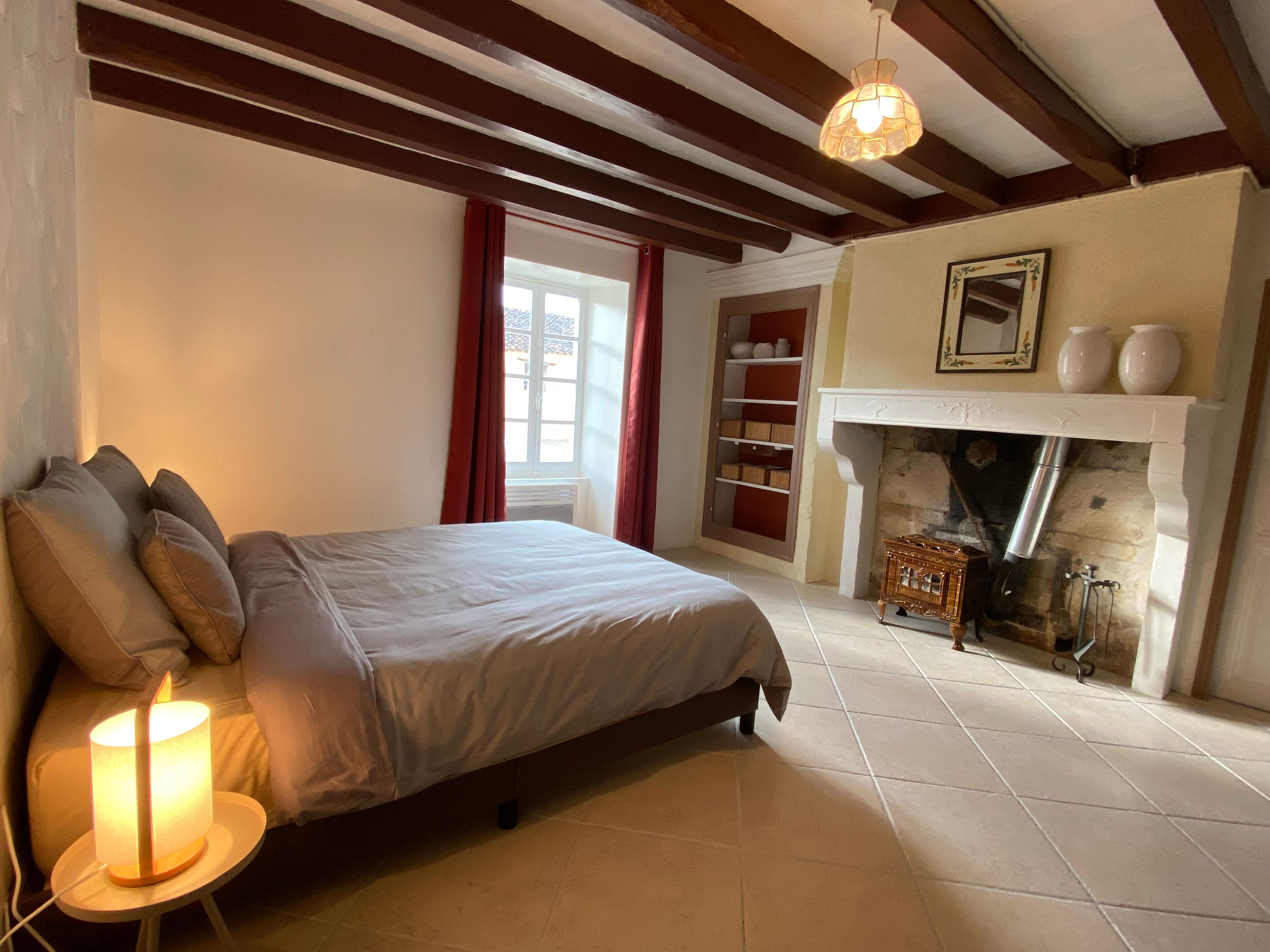 Un dormitorio con cama, mesita de noche con lámpara, chimenea y ventana con cortinas.
