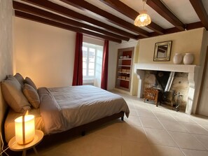 Un dormitorio con cama, mesita de noche con lámpara, chimenea y ventana con cortinas.