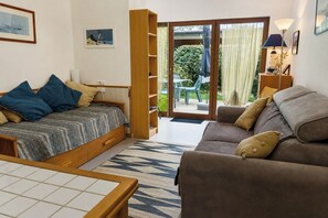 A room with a bed, a sofa, a table, and a sliding glass door leading to an outdoor area with a patio and greenery. - Beachfront studio in a gated community, direct beach access, private parking (La Turballe)
