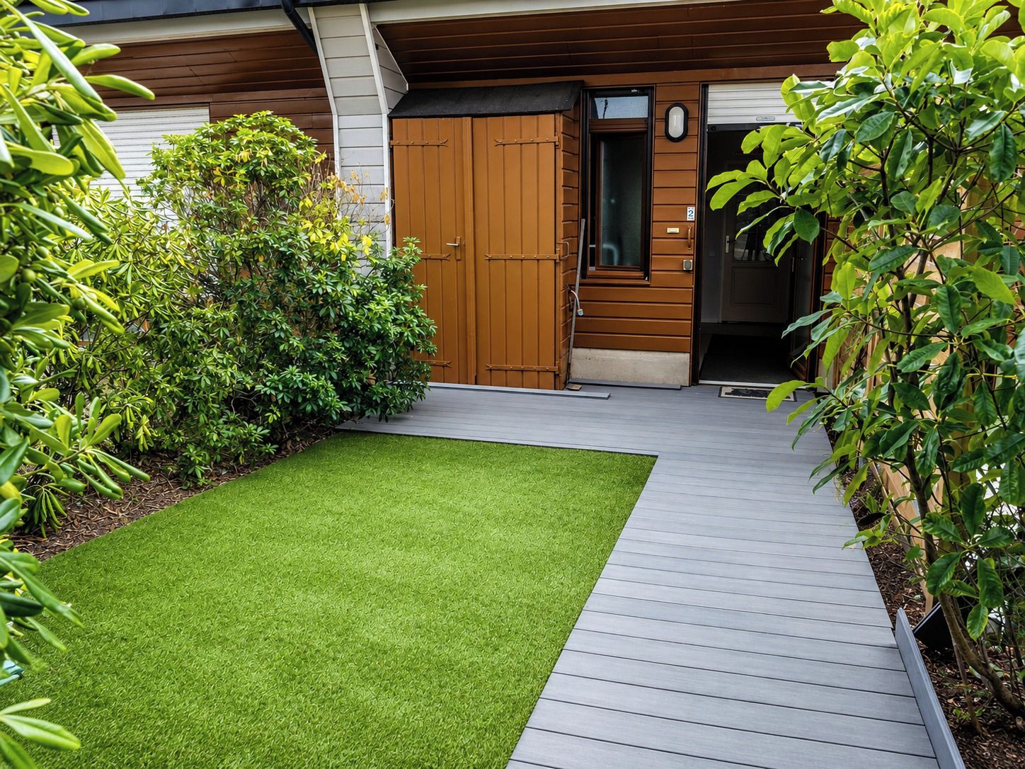 A modern building with a wooden door, a small garden with artificial grass, and a wooden deck.