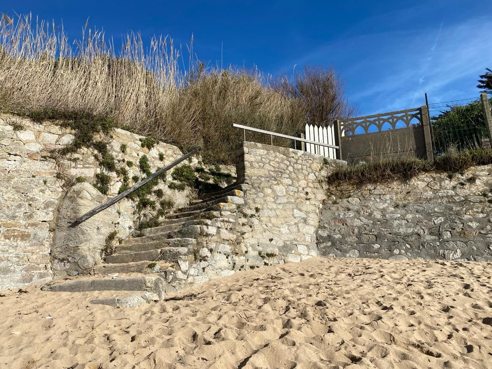 A stone staircase leading down to a sandy area with a white fence and vegetation.