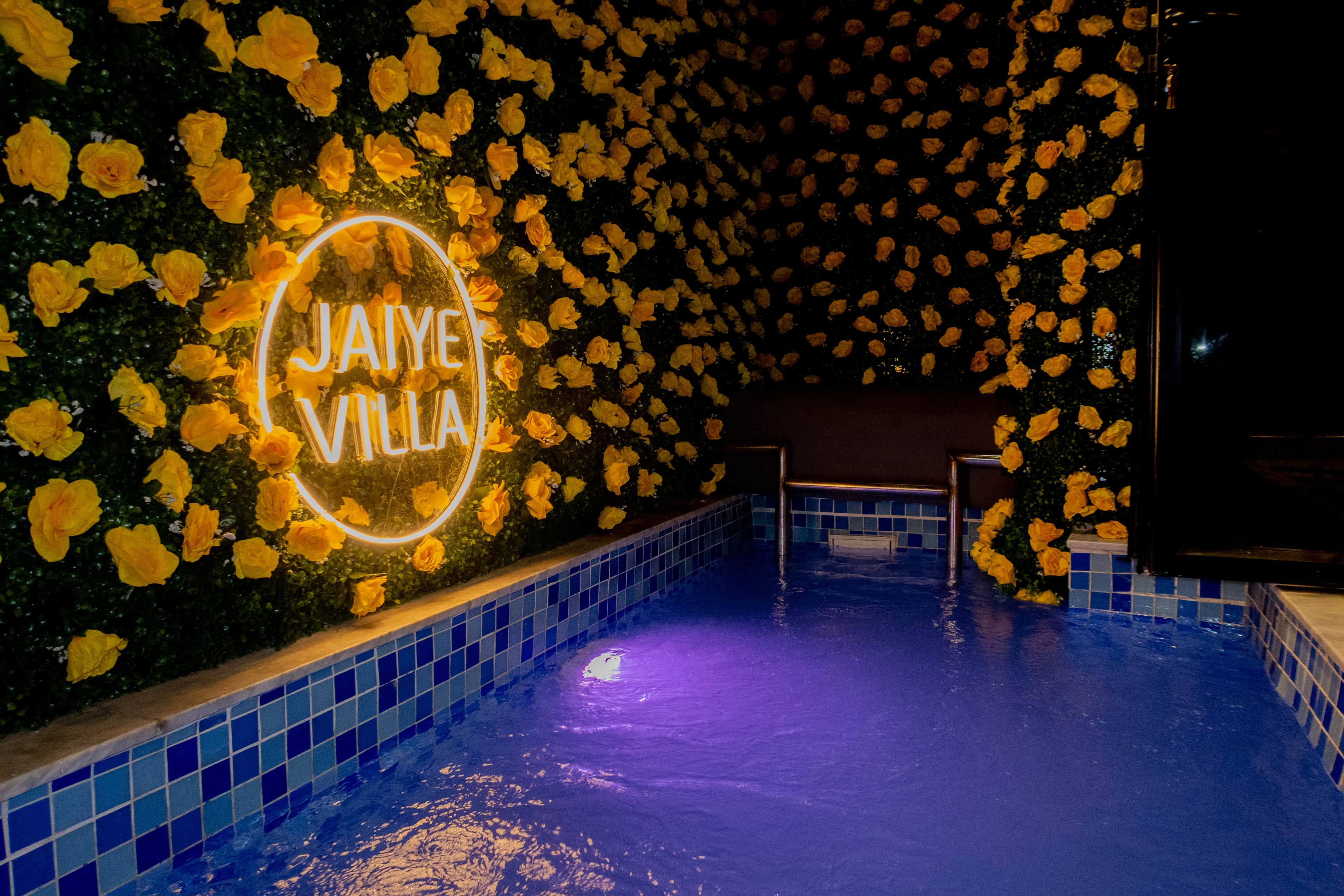 A swimming pool area with a neon sign reading 'Jaiye Villa' surrounded by yellow flowers.