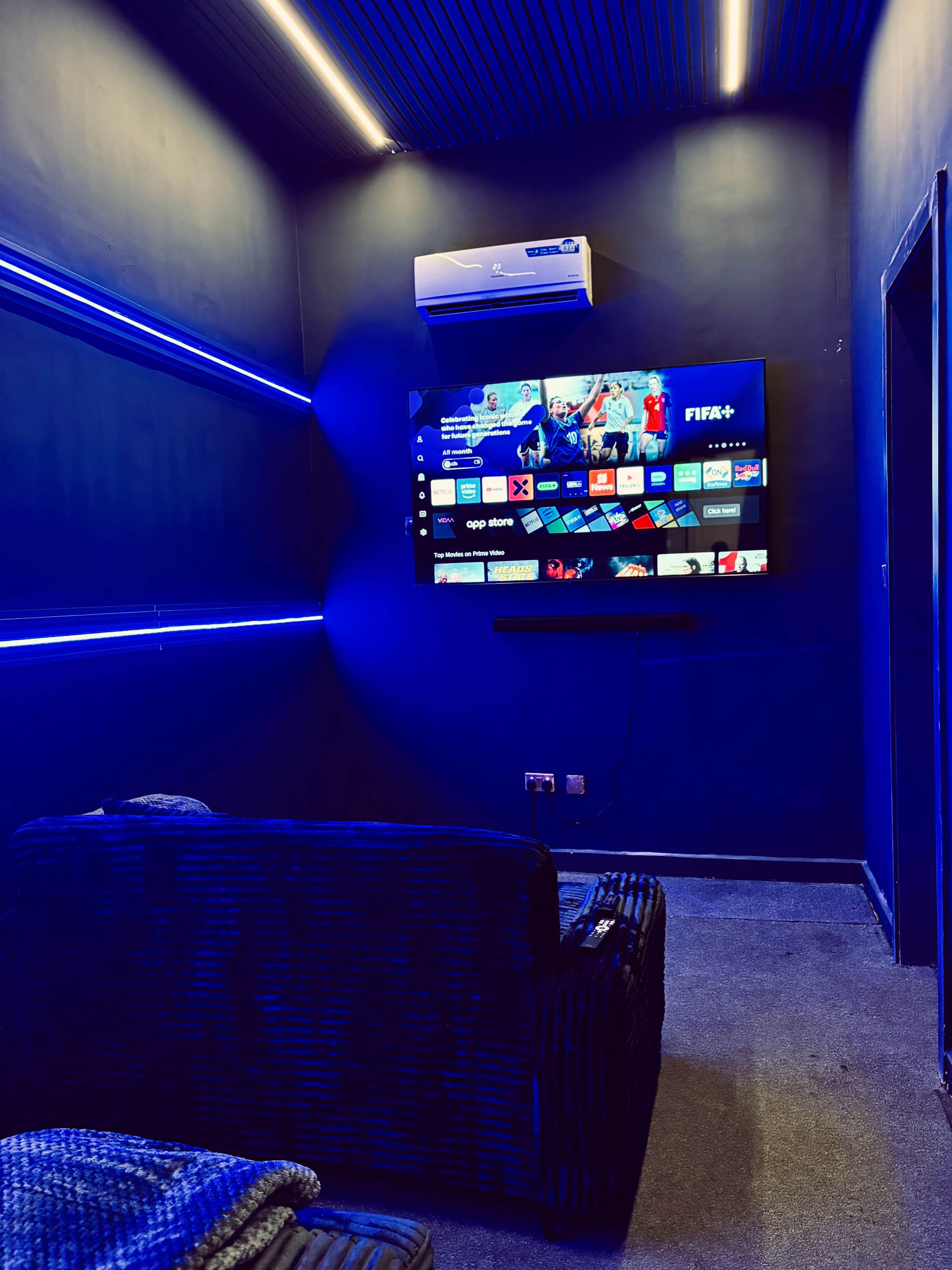 A room with a flat-screen TV mounted on the wall, a comfortable seating area with a sofa, and an air conditioning unit above the TV.