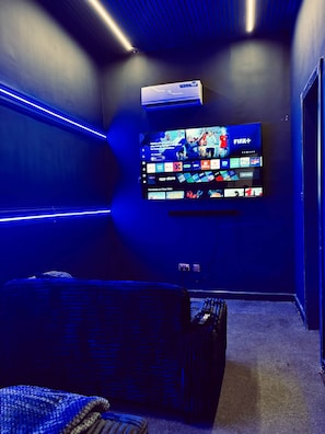 A room with a flat-screen TV mounted on the wall, a comfortable seating area with a sofa, and an air conditioning unit above the TV. - Jaiye Villa Entire Place (Lekki)