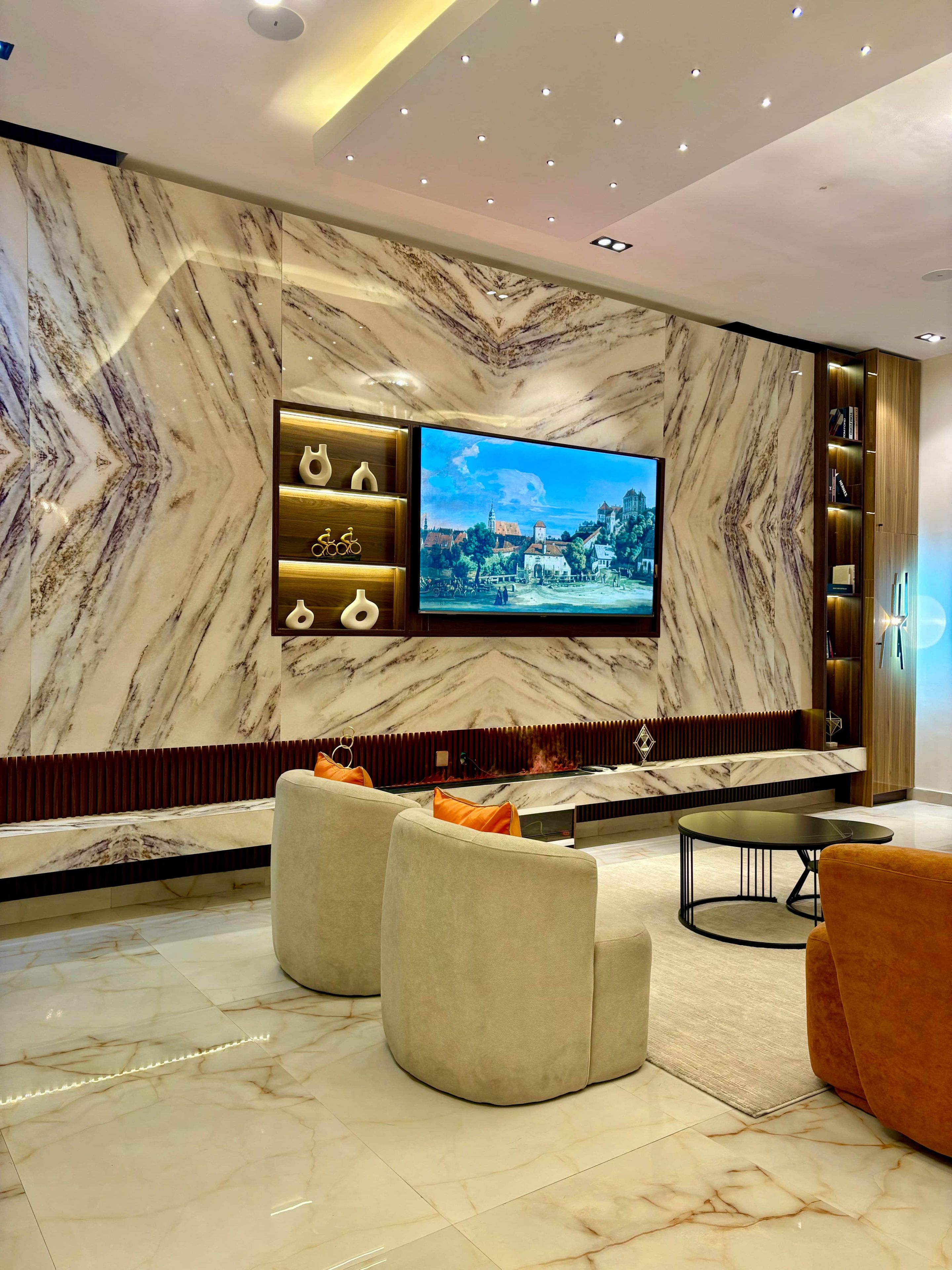A modern living room with a large flat-screen TV mounted on a marble wall, two sofas, a coffee table, and a bookshelf with decorative items.