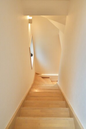 A staircase with wooden steps leading upwards, white walls, and a light fixture on the left. - A 6-minute walk from Shinjuku-Shin-Okubo Station! 4-Bedroom, 3-Story House for Exclusive Rental (Sleeps Up to 8) | 15-Minute Walk from Kabukicho (東京都新宿区大久保二丁目)